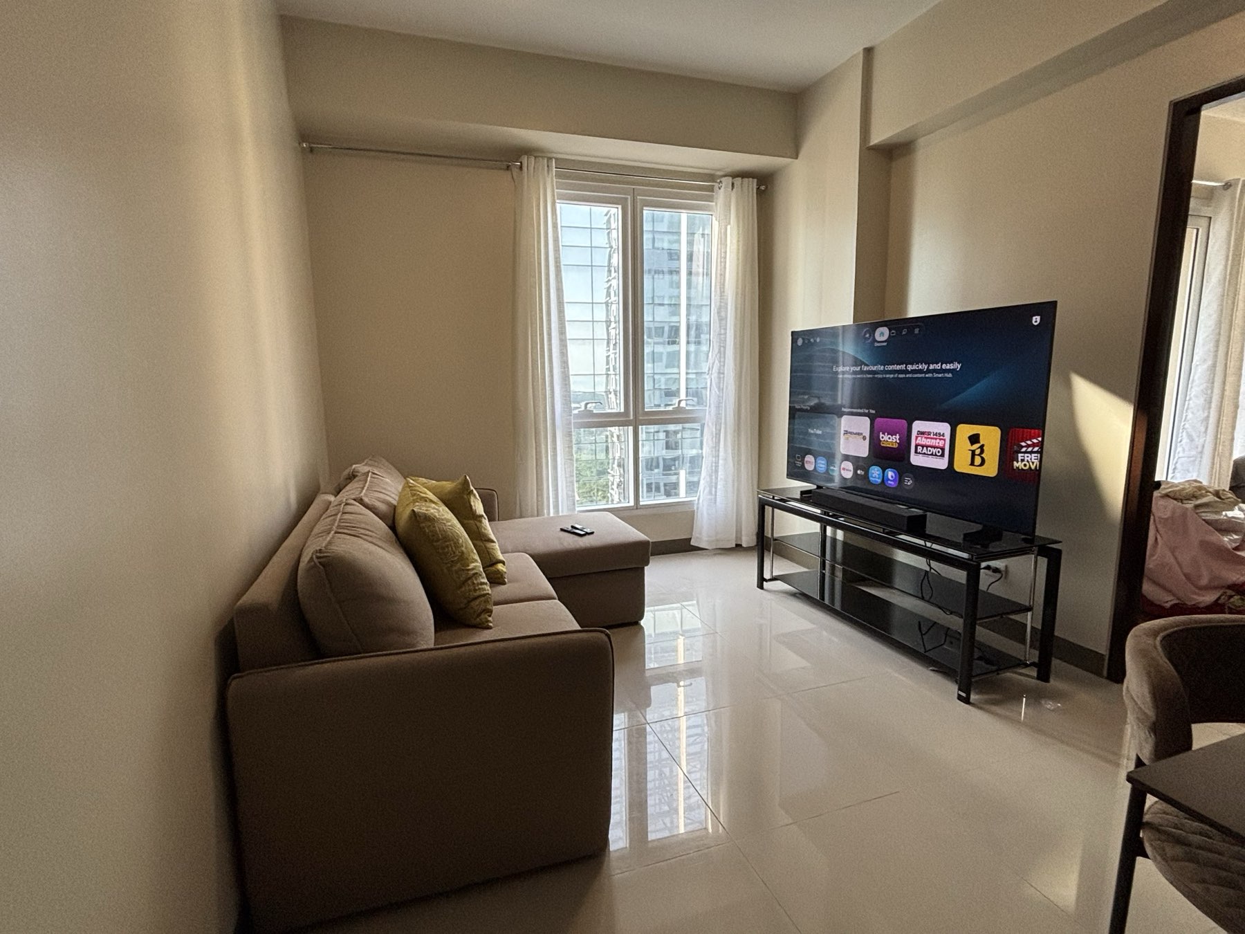 Open-plan living and dining room in the DAYON TA two-bedroom condo at Mactan Newtown, with a beige sectional sofa, large smart TV, and tower views through a curtained window