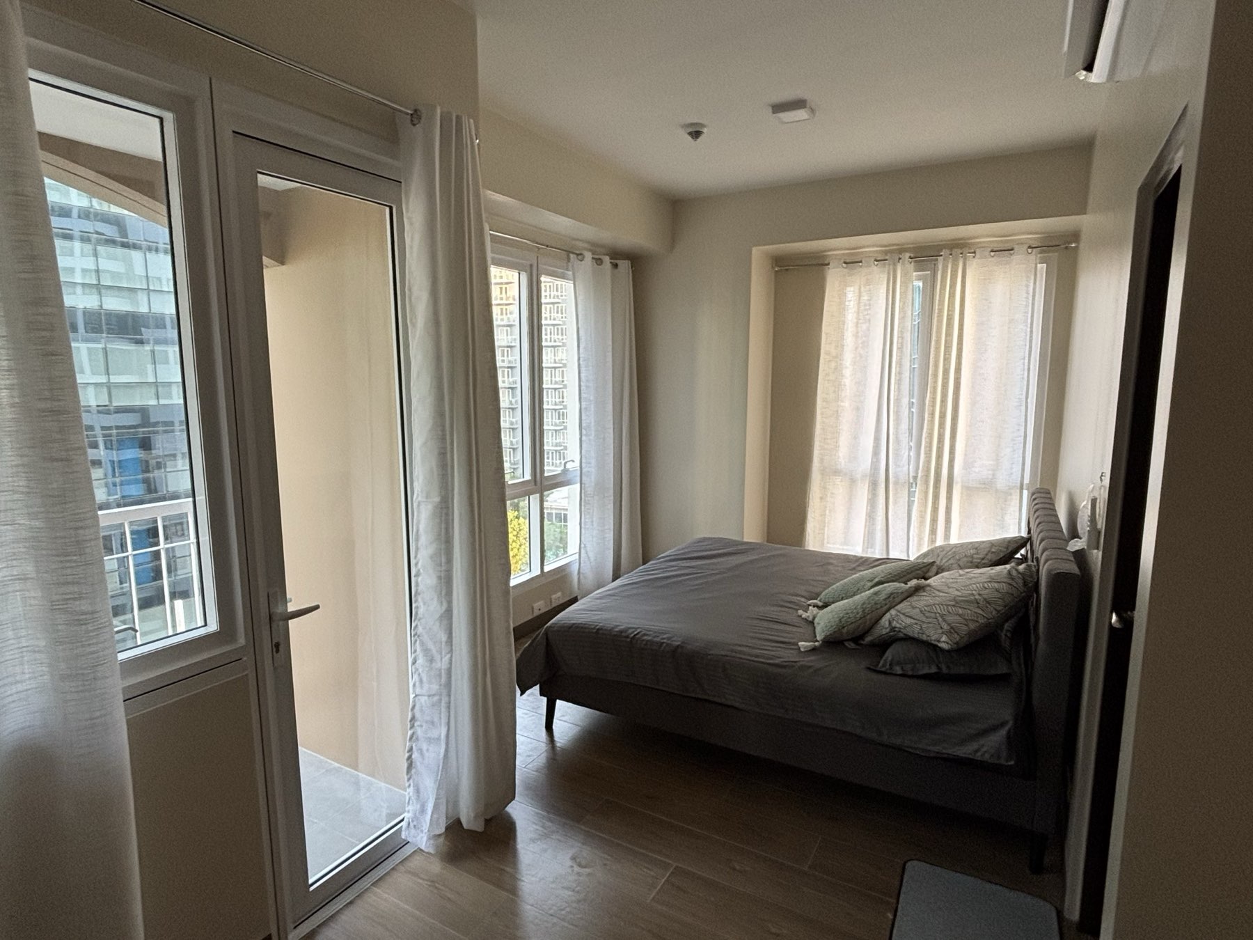Primary bedroom of the DAYON TA condo, with a queen bed, wood floors, and a corner balcony door letting in daylight over Mactan Newtown